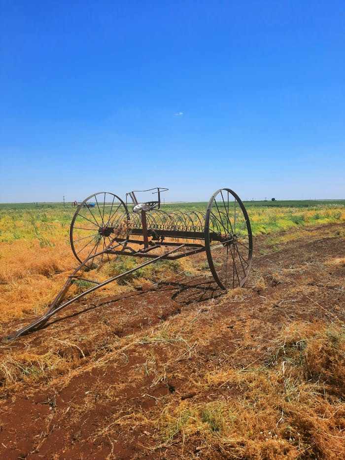 Vintage farm rake rusting in a vast rural field under clear blue skies.